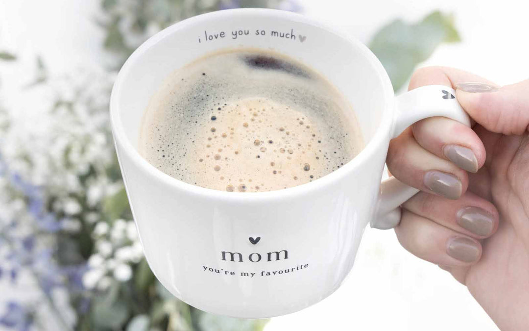 Eine Hand hält vor einem floralen Hintergrund eine weiße Tasse mit der Aufschrift „Mama, ich liebe dich so sehr, du bist mein Liebling“, gefüllt mit Kaffee.