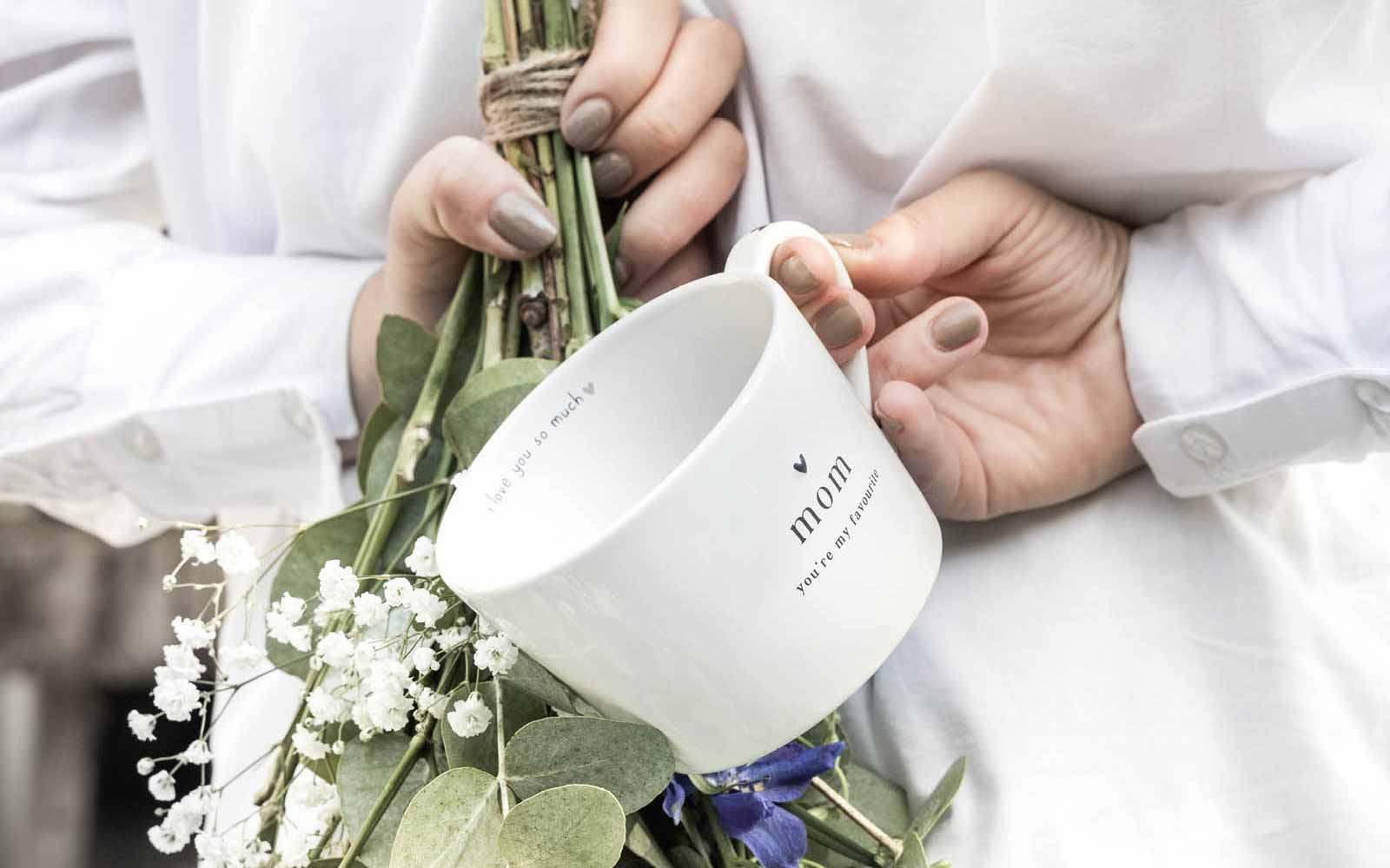 Nahaufnahme von Händen, die eine weiße Tasse mit der Aufschrift „Mama“ und einem Blumenstrauß halten, als Symbol für ein Geschenk oder eine Ehrerbietung.