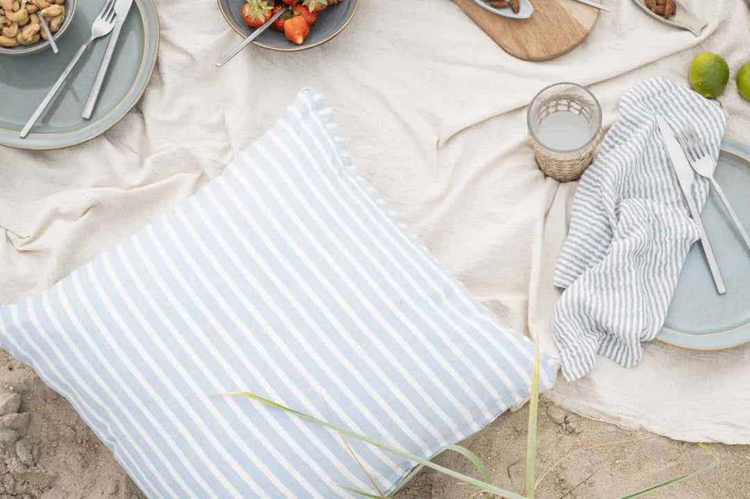 Ein Strandpicknick-Set bestehend aus einem gestreiften Kissen, einer Decke, Tellern mit Essen, Besteck und einer Kerze auf einer sandigen Oberfläche.
