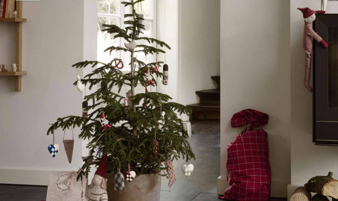 Ein kleiner, mit einfachen Ornamenten geschmückter Weihnachtsbaum steht in einem Topf neben einer Treppe. Zwei Stofftiere und ein karierter Sack liegen in der Nähe auf dem Boden.