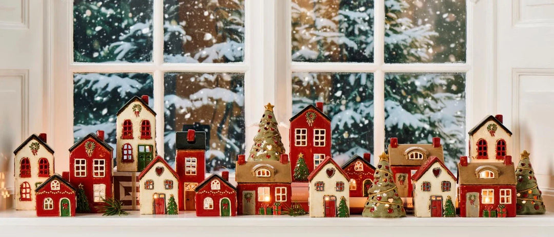 Auf einer Fensterbank sind Miniaturweihnachtshäuser und Weihnachtsbäume ausgestellt, und außerhalb des Fensters sind schneebedeckte Bäume zu sehen.