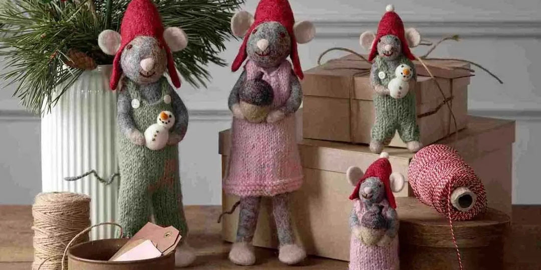 Vier Filzmausfiguren mit roten Hüten stehen auf einem Tisch mit Geschenkkartons, einer Spule mit roter Schnur und einer Vase mit Grünzeug im Hintergrund.