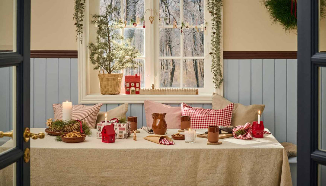 Ein gemütlicher Essbereich mit einem weihnachtlich gedeckten Tisch. Zur Dekoration gehören Kerzen, ein kleiner Baum, rote und weiße Akzente, Kissen und ein Blick durch ein Fenster auf kahle Bäume.