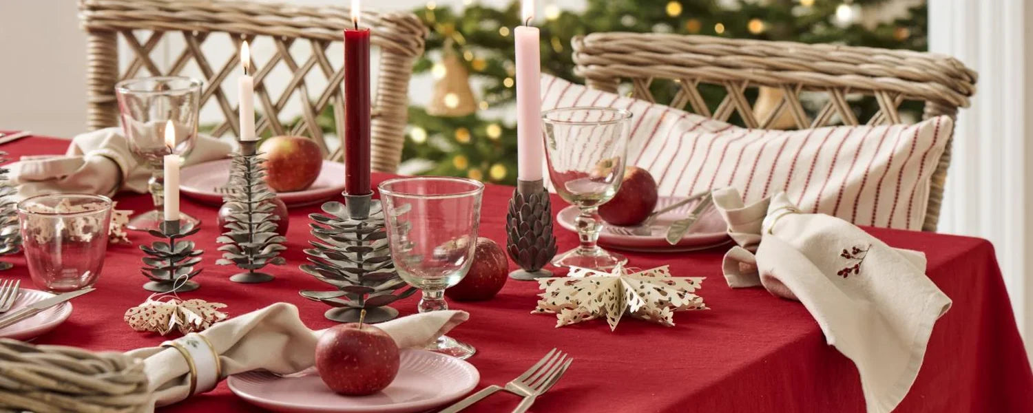 Ein festlich gedeckter Tisch mit roter Tischdecke, Kerzenhaltern aus Metall, Äpfeln, Gläsern und gefalteten Servietten vor einem Weihnachtsbaum mit Lichtern.