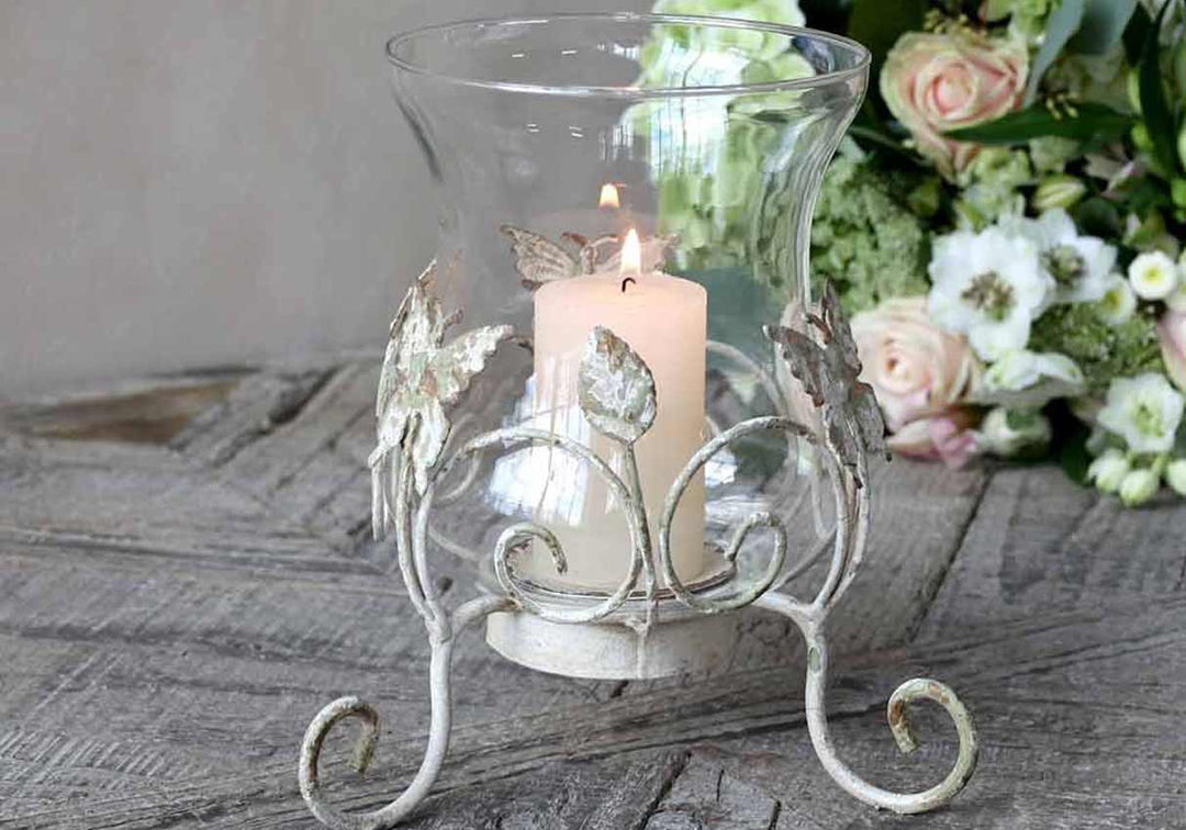 Eine brennende Kerze in einem dekorativen Glashalter mit Metallblatt-Akzenten auf einer Holzoberfläche, neben einem Blumenarrangement.