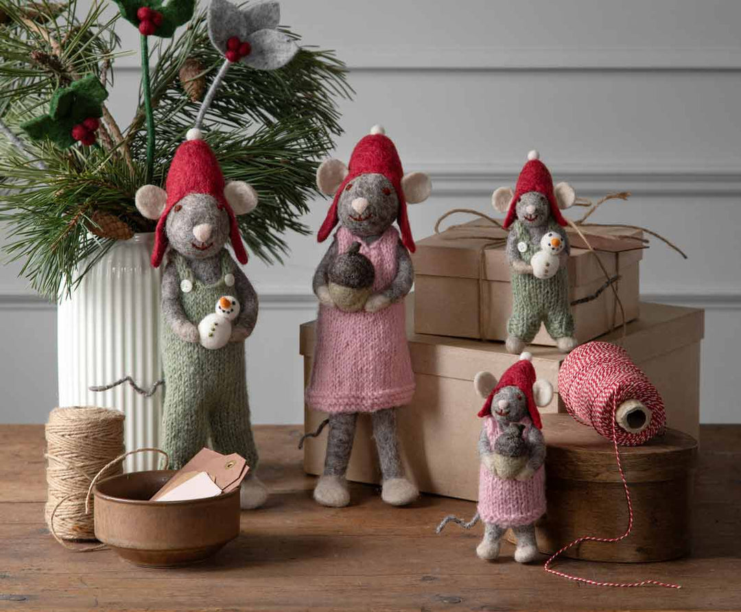 Vier Filzmäuse in Strickkostümen stehen neben Geschenkboxen und einem Weihnachtsbaum, mit einer Schüssel und Garn auf einem Holztisch.