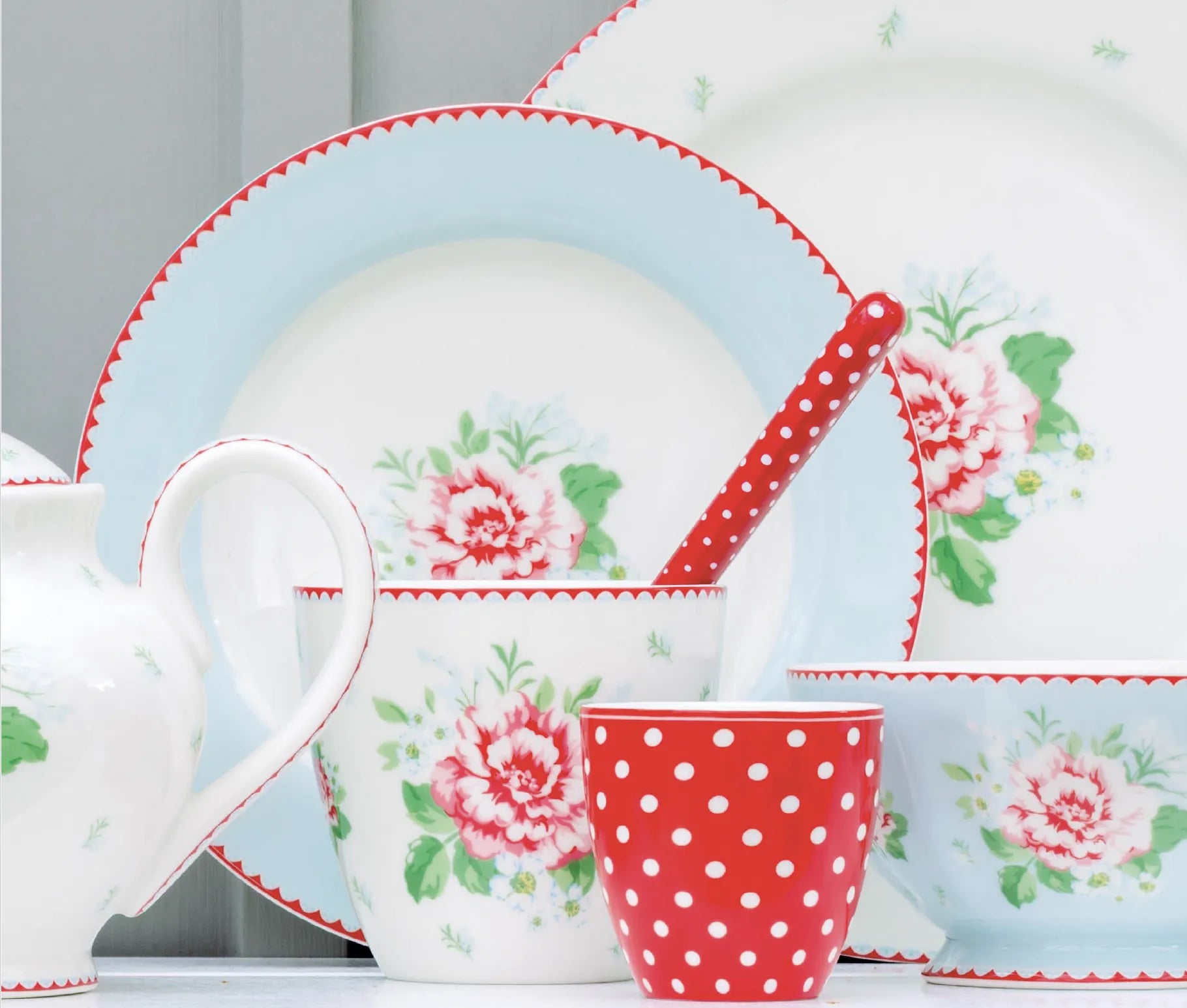Verschiedene Keramikteller mit floralen Mustern und pastellblauen Rändern, eine rot-weiß gepunktete Tasse und ein passender Löffel sind zusammen angeordnet.