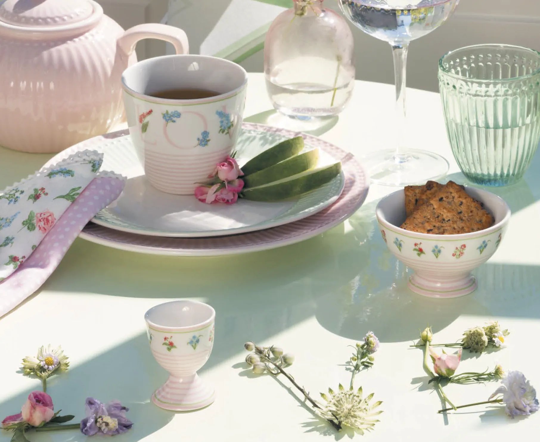 Ein gedeckter Tisch mit einer Teetasse, einer Untertasse, Crackern in einer kleinen Schale, einem Glas, einer Serviette und verschiedenen frischen Blumen, die auf einer hellen Fläche angeordnet sind.
