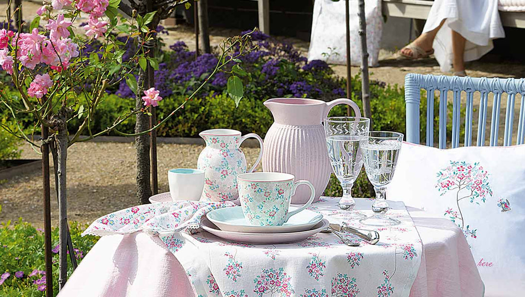 Ein Gartentisch mit Tellern, Tassen, einem Krug, Glaswaren und Servietten mit Blumenmuster, umgeben von blühenden Blumen und Grünpflanzen.