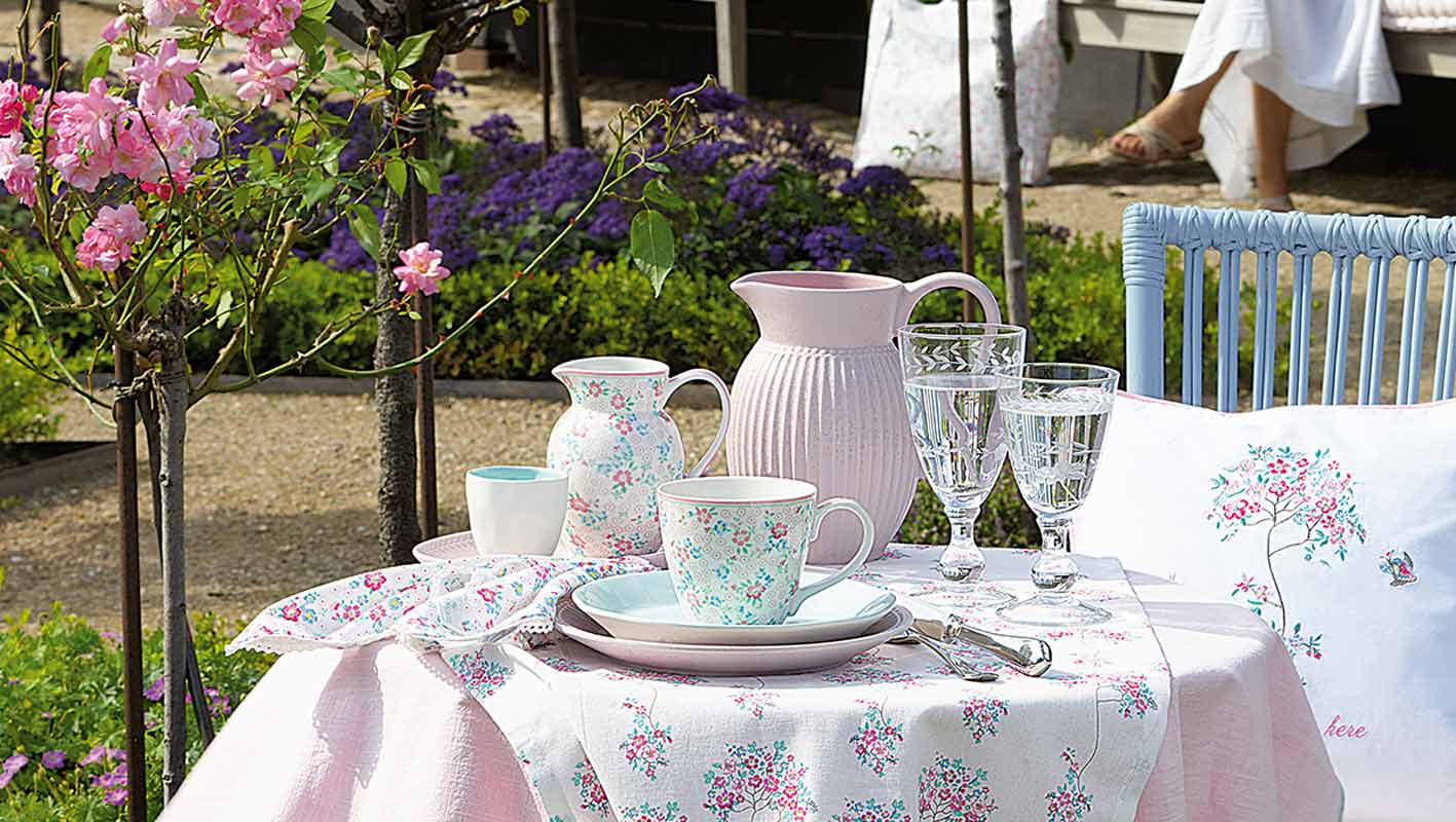 Ein Gartentisch mit Tellern, Tassen, einem Krug, Glaswaren und Servietten mit Blumenmuster, umgeben von blühenden Blumen und Grünpflanzen.