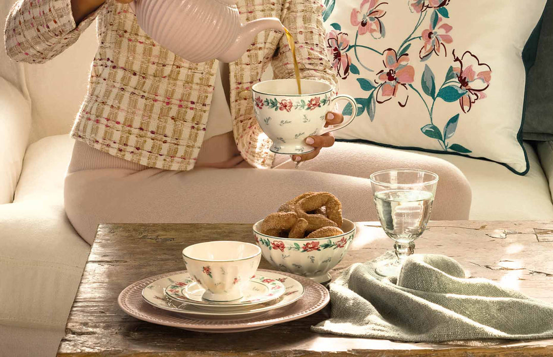 Eine Person gießt Tee aus einer Teekanne in eine Tasse mit Blumenmuster auf einem Holztisch. Auf dem Tisch steht verschiedenes Geschirr, ein Glas Wasser, eine Stoffserviette und eine Schüssel mit Gebäck.