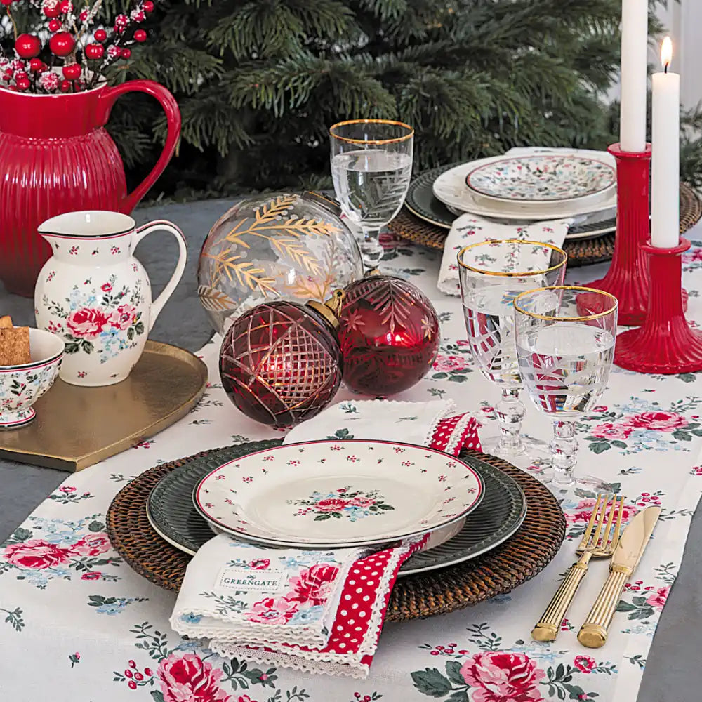 Eine festlich gedeckte Tafel mit Tellern mit Blumenmuster, Gläsern, Kerzen, rot-goldenen Dekorationselementen und einem Tischläufer mit Blumenmuster, die in der Nähe eines Weihnachtsbaums steht.