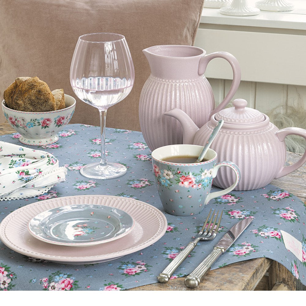 Ein pastellrosa- und pastellblaues Teeservice mit Blumenmuster, bestehend aus einer Teekanne, einer Tasse, einer Untertasse, einem Krug, einem Wasserglas, einem Brötchen und Besteck, angerichtet auf einer blauen Tischdecke mit Blumenmuster.