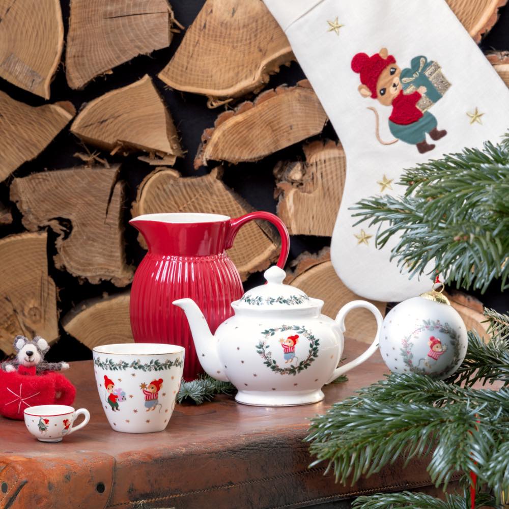 Ein festliches Teeservice mit Tassen, Teekanne und rotem Krug ist neben einem Weihnachtsstrumpf und einem Weihnachtsbaum arrangiert, im Hintergrund ist gestapeltes Brennholz zu sehen.
