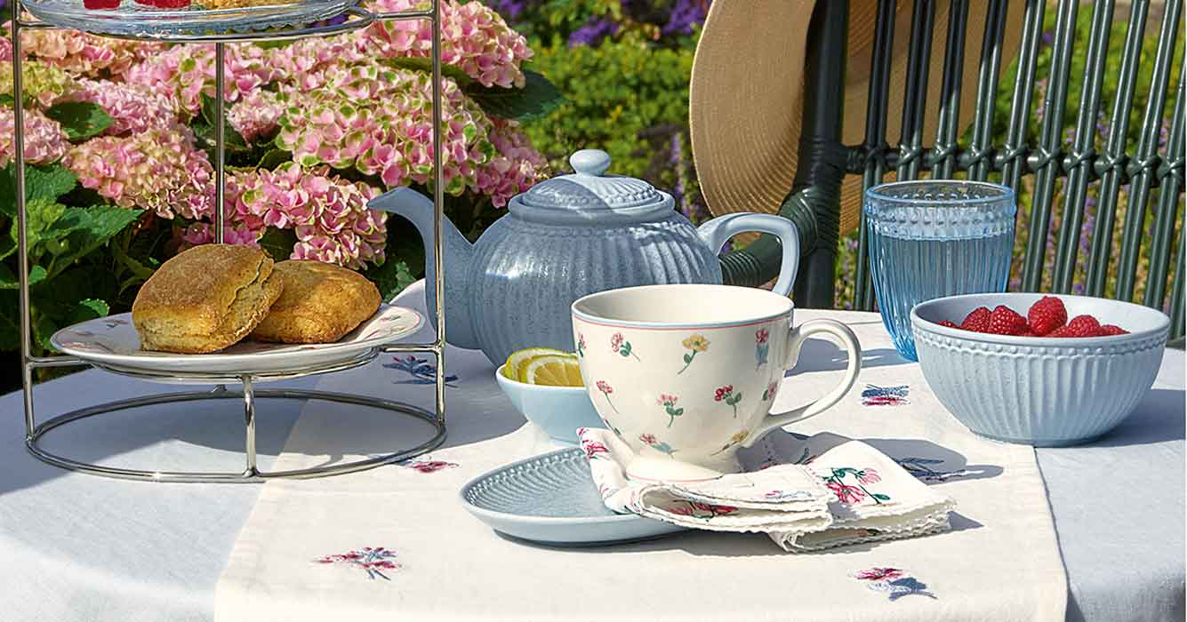 Ein für den Tee gedeckter Tisch mit einer Teekanne, einer Teetasse mit Blumenmuster, einer Schale mit Himbeeren, Scones auf einem Ständer, einem Glas Wasser und Blumen im Hintergrund.
