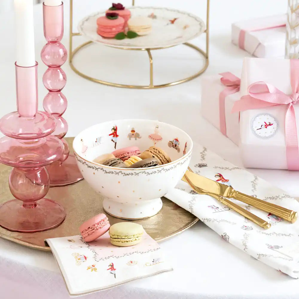Eine Schale und ein Teller mit bunten Macarons auf einem Tisch, der mit rosa Glaskerzenhaltern, goldenem Besteck, einer Geschenkbox mit einer rosa Schleife und dekorativen Servietten gedeckt ist.