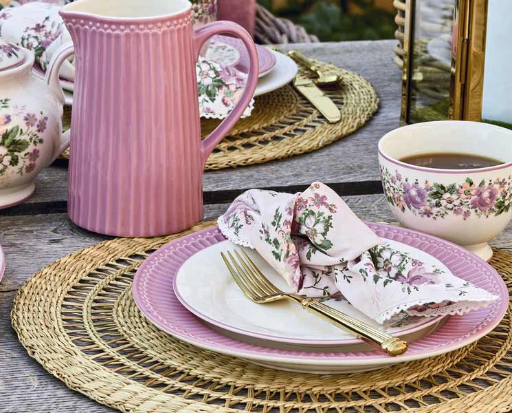 Tischgedeck für den Außenbereich mit rosa Geschirr, Servietten mit Blumenmuster und einer Kanne, arrangiert auf gewebten Tischsets mit einer Tasse Tee.