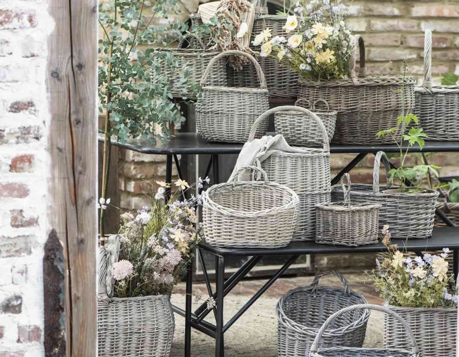 Weidenkörbe mit verschiedenen Trockenblumen, ausgestellt auf einem Metallregal vor einer Backsteinwand in rustikalem Ambiente.