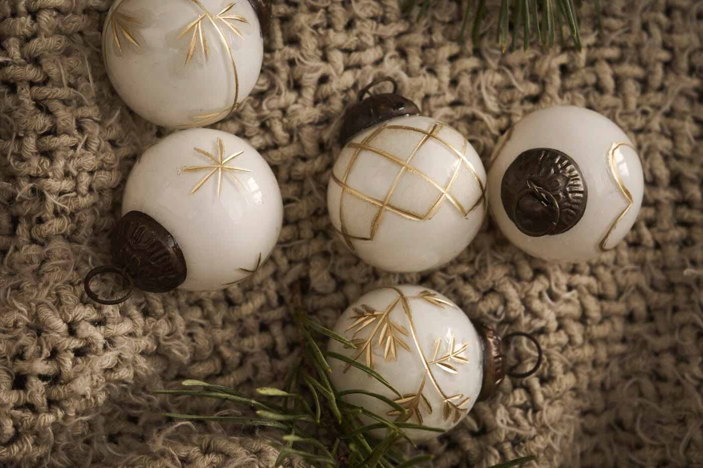 Fünf weiße Weihnachtsornamente mit goldenem Muster ruhen auf einem beigen Strickstoff, begleitet von grünen Tannennadeln.
