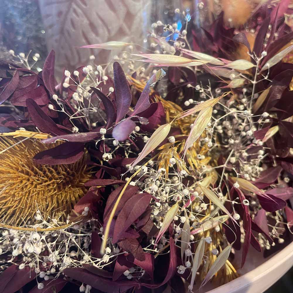 Eine Nahaufnahme des Alsaba Home - Deko Kranz Herbst mit getrockneten violetten Blättern, weißem Schleierkraut, beigen Gräsern und einer stacheligen goldenen Samenkapsel.