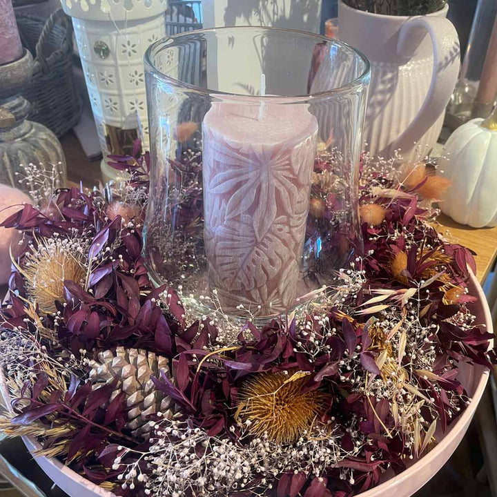 Die Alsaba Home - Deko Kranz Herbst zeigt eine rosafarbene Kerze in einem Glashalter, umgeben von getrockneten lila Blättern, Tannenzapfen und kleinen Blumen, arrangiert auf einem Holztisch.