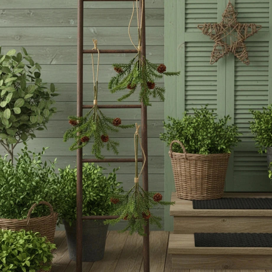 Ein Alsaba Home Künstlicher Tannenzweig mit Zapfen 28 cm schmückt eine Leiter neben grünen Topfpflanzen auf einer hölzernen Veranda, unter einem sternförmigen Kranz an der Wand.