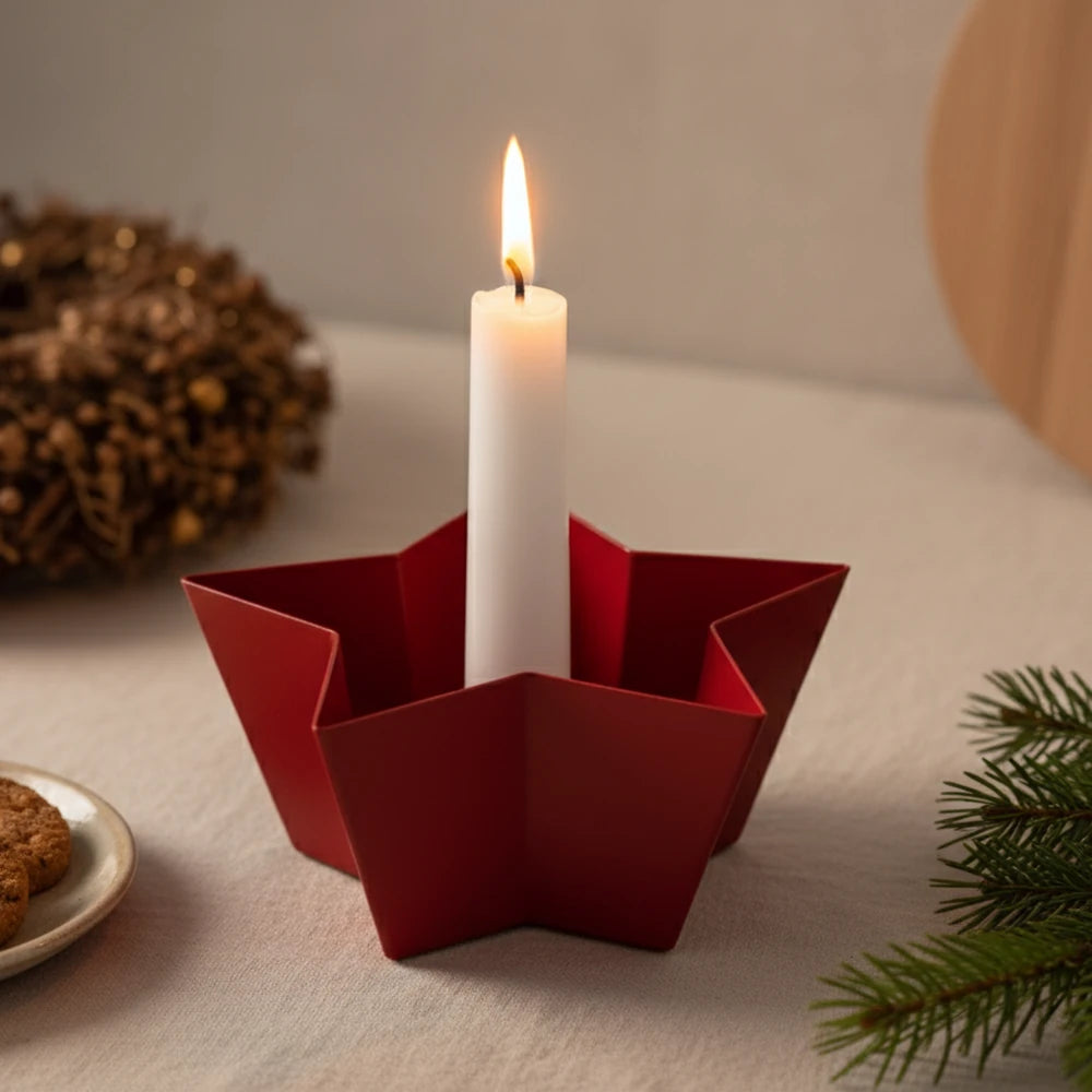 Eine brennende weiße Kerze steht in einem roten, sternförmigen Metallhalter von Alsaba Home auf einem Tisch mit einem Kranz, Keksen und Tannenzweigen im Hintergrund.