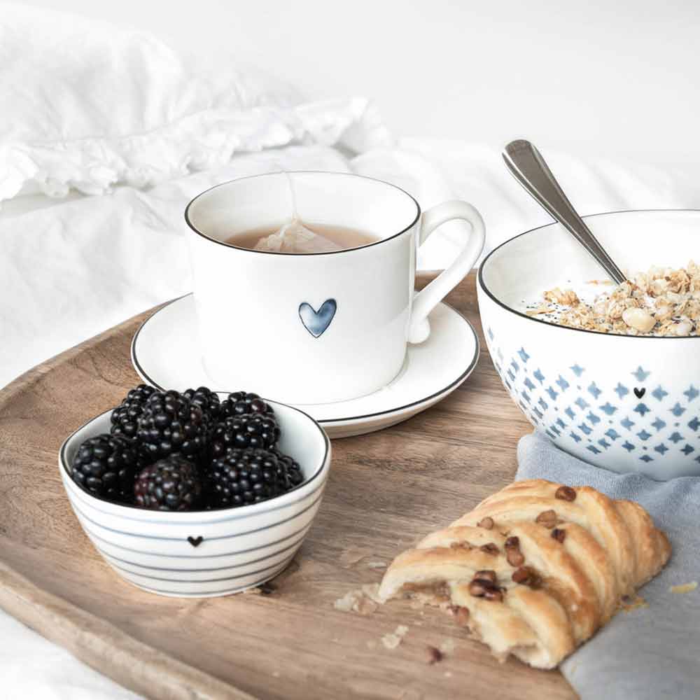 Eine ruhige Frühstücksszene mit einer Bastion Collection – Tasse Iris Blue Heart, einer Schüssel Müsli, Brombeeren und Gebäck auf einem hölzernen Serviertablett.