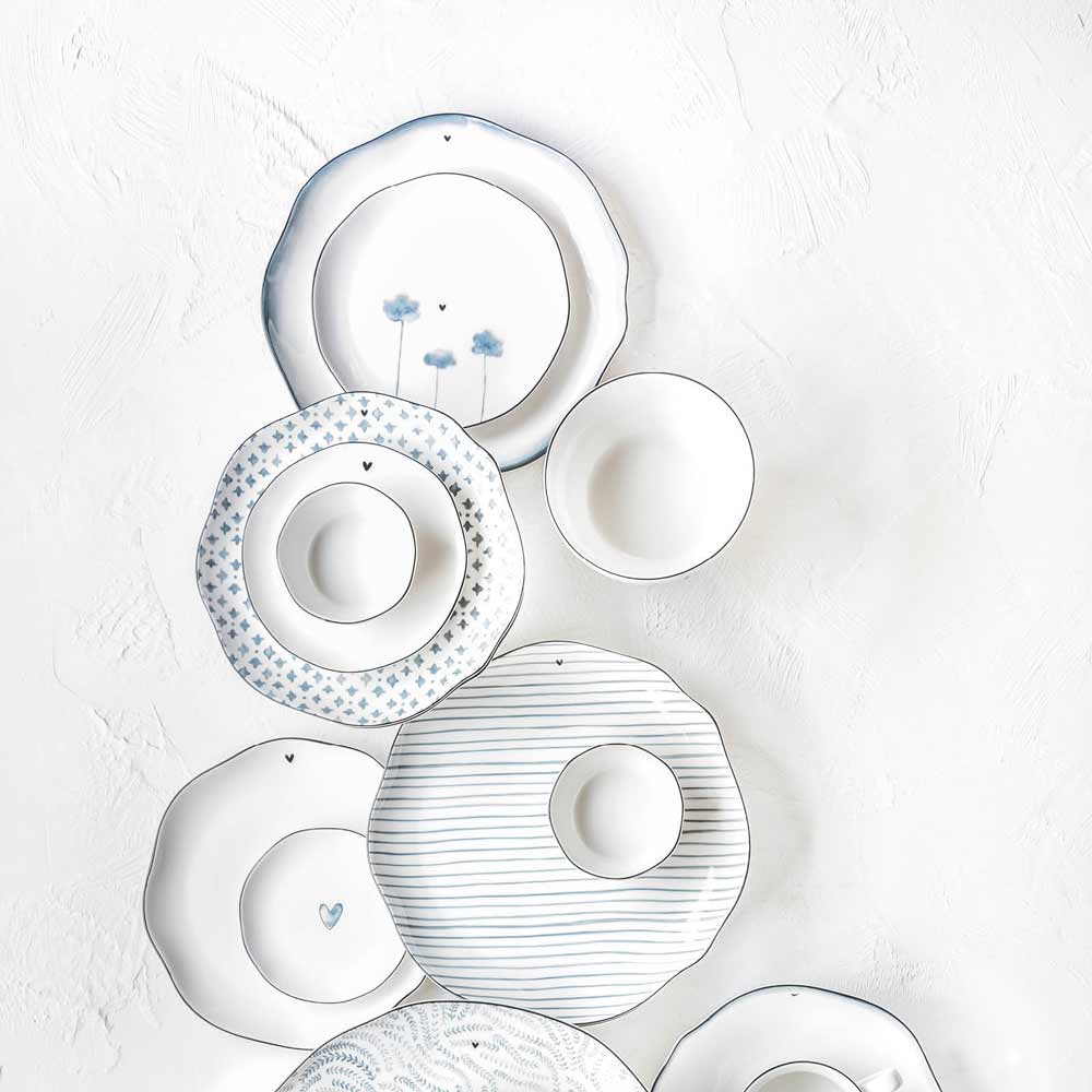 Die Bastion Collections Iris Blue Streifen umfassen weiße Porzellanteller und -schüsseln mit blauen dekorativen Mustern, die in einem Halbkreis auf einer strukturierten weißen Oberfläche angeordnet sind.
