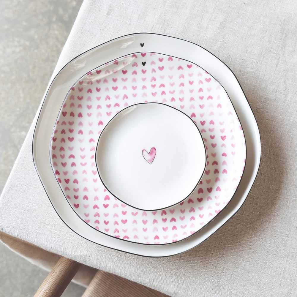 Zwei Bastion Collections Frühstücksteller Rose Hearts all over sind auf einer beigen Tischdecke gestapelt; der kleinere Teller zeigt ein einzelnes rosa Herz in der Mitte.