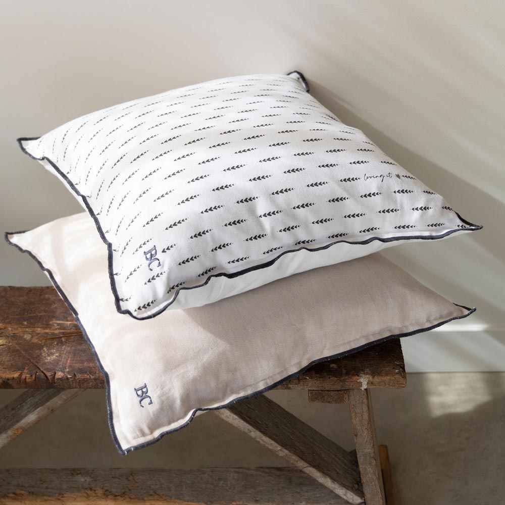 Auf einer rustikalen Holzbank liegen zwei Kissen: Das obere ist ein Kissenbezug „Chambray Hearts“ von Bastion Collections in Weiß mit schwarzem Muster und Text, während das untere schlicht beige ist.