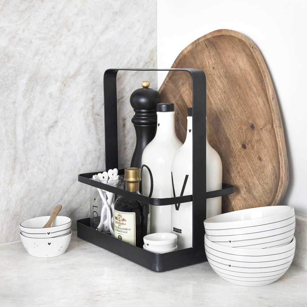 Eine Küchenarbeitsplatte mit einem Bastion Collections - Küchen Organizer Metall Schwarz, auf dem weiße Flaschen, eine Pfeffermühle und Utensilien stehen. Hinter dem Organizer befindet sich ein hölzernes Schneidebrett. Davor stehen zwei weiße Schüsseln mit Streifen.