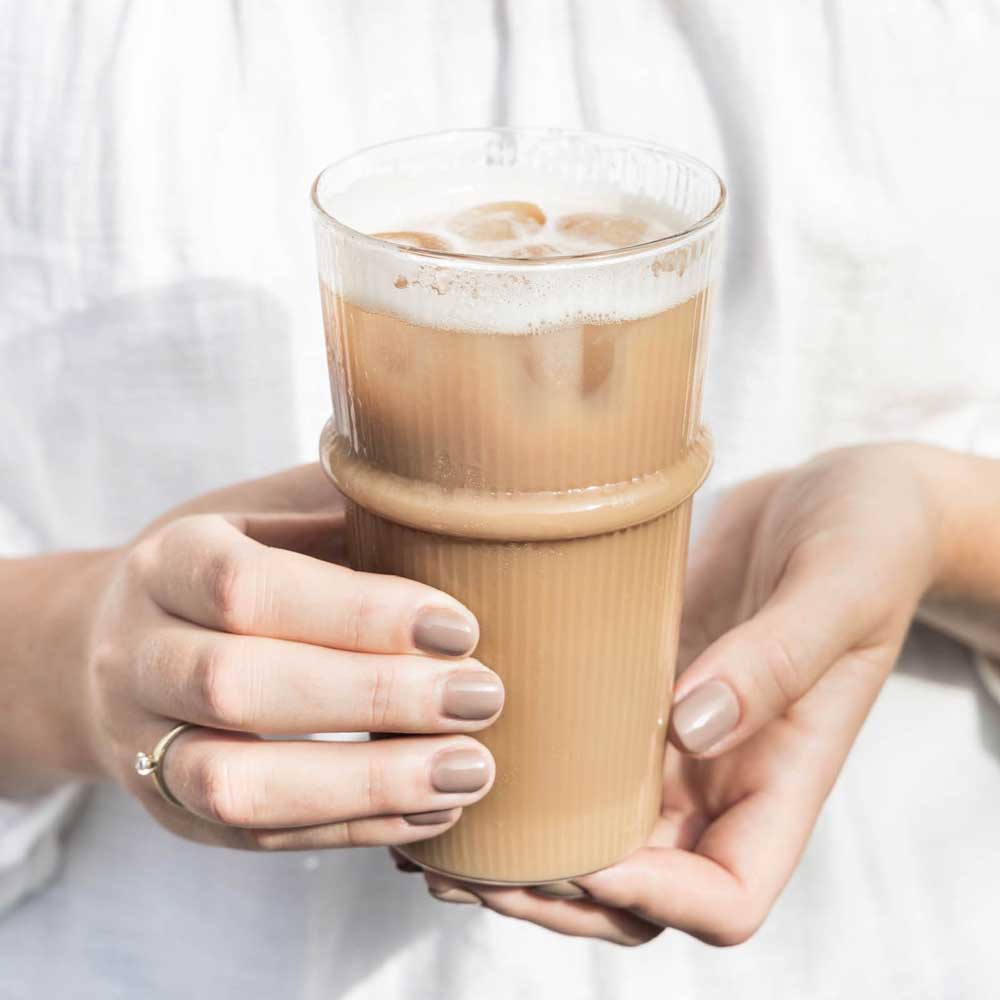 Eine Person hält mit beiden Händen vor einem hellen Hintergrund ein mit Eiskaffee gefülltes Bastion Collections - Latte Macchiato Glas Riffel.