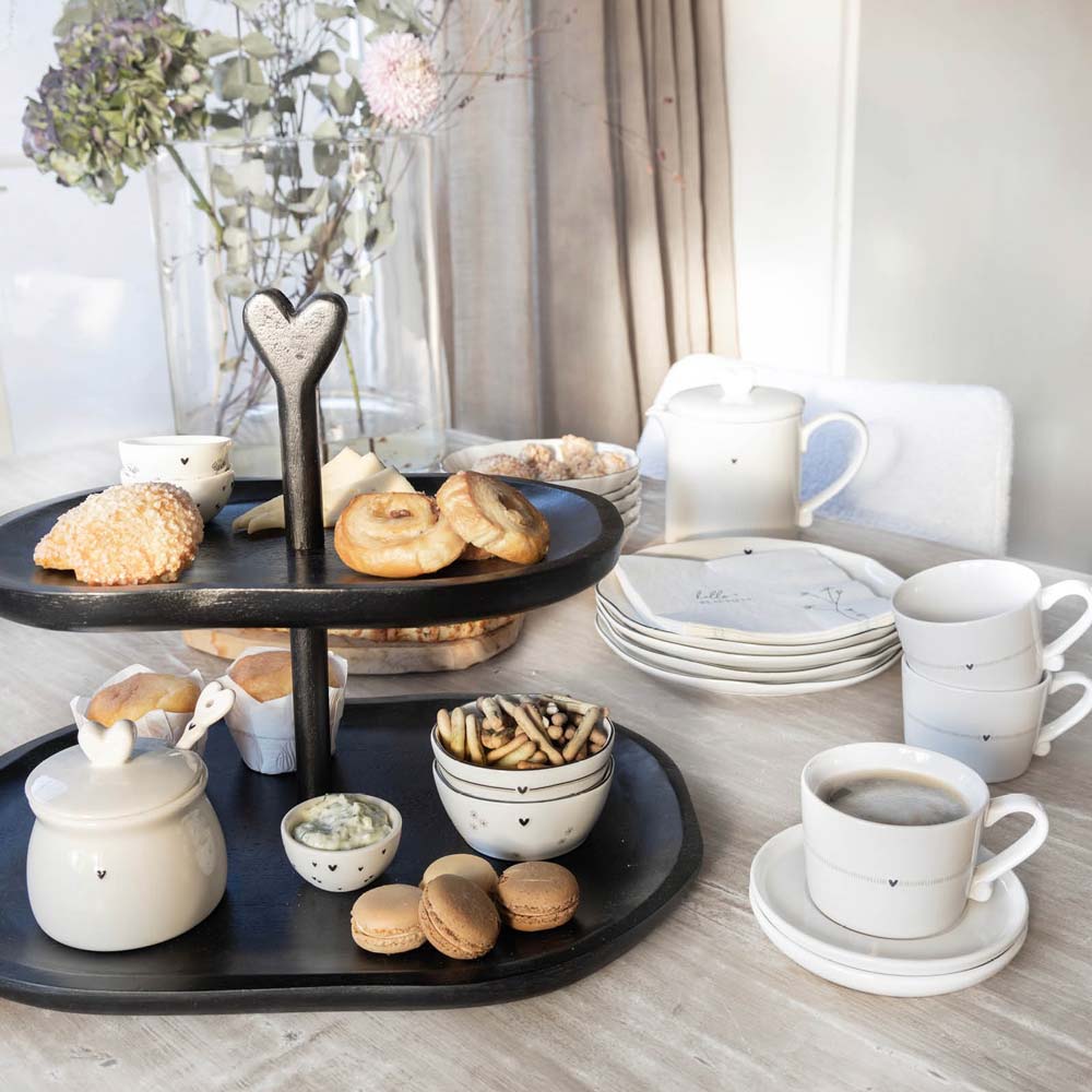 Ein zweistöckiges Serviertablett mit Bastion Collections - Marmeladendose mit Löffel Herz, Keksen und einer Tasse Kaffee auf einem Tisch, umgeben von Tassen und Tellern.