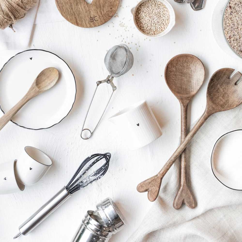 Verschiedene Küchenutensilien und Geschirr, darunter ein Salatbesteck Holz Natur Set von Bastion Collections, ein Schneebesen, Tassen, Schüsseln und ein Tee-Ei, sind zusammen mit verschüttetem Getreide und Mehl auf einer weißen Oberfläche angeordnet.