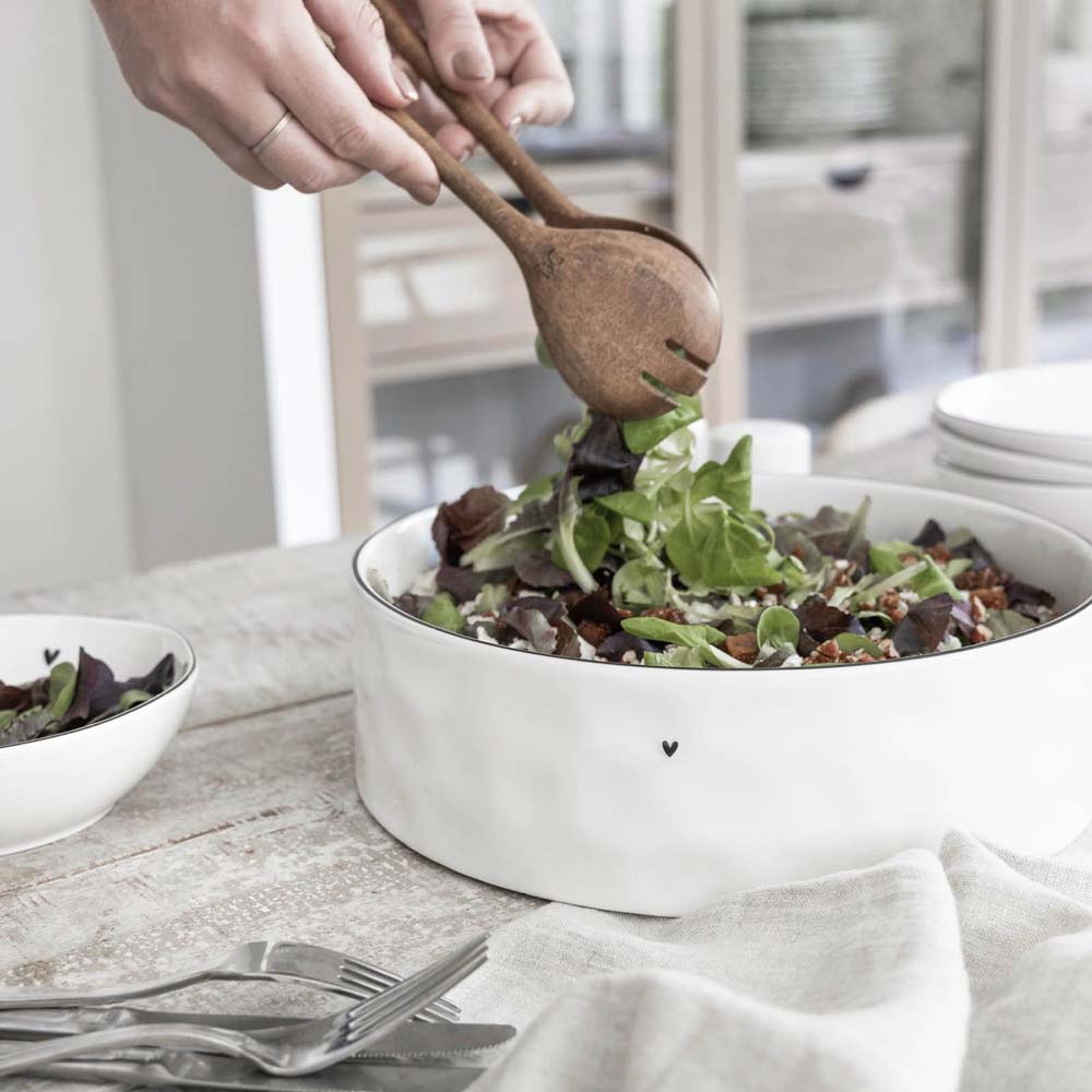 Eine Person rührt einen Salat mit Blattgemüse und Gemüse mit einem hölzernen Salatbesteck in einer weißen Salatschüssel in Herzform von Bastion Collections auf einem mit Besteck und Servietten gedeckten Tisch.