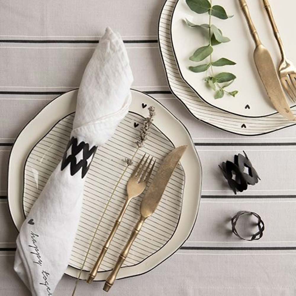 Ein gedeckter Tisch besteht aus weißen Tellern mit Blattakzenten und Herzen, goldenem Besteck und einem Serviettenring „Herzen“ von Bastion Collections. Das goldene Messer und die goldene Gabel liegen neben einer gefalteten Serviette auf einer gestreiften Tischdecke.