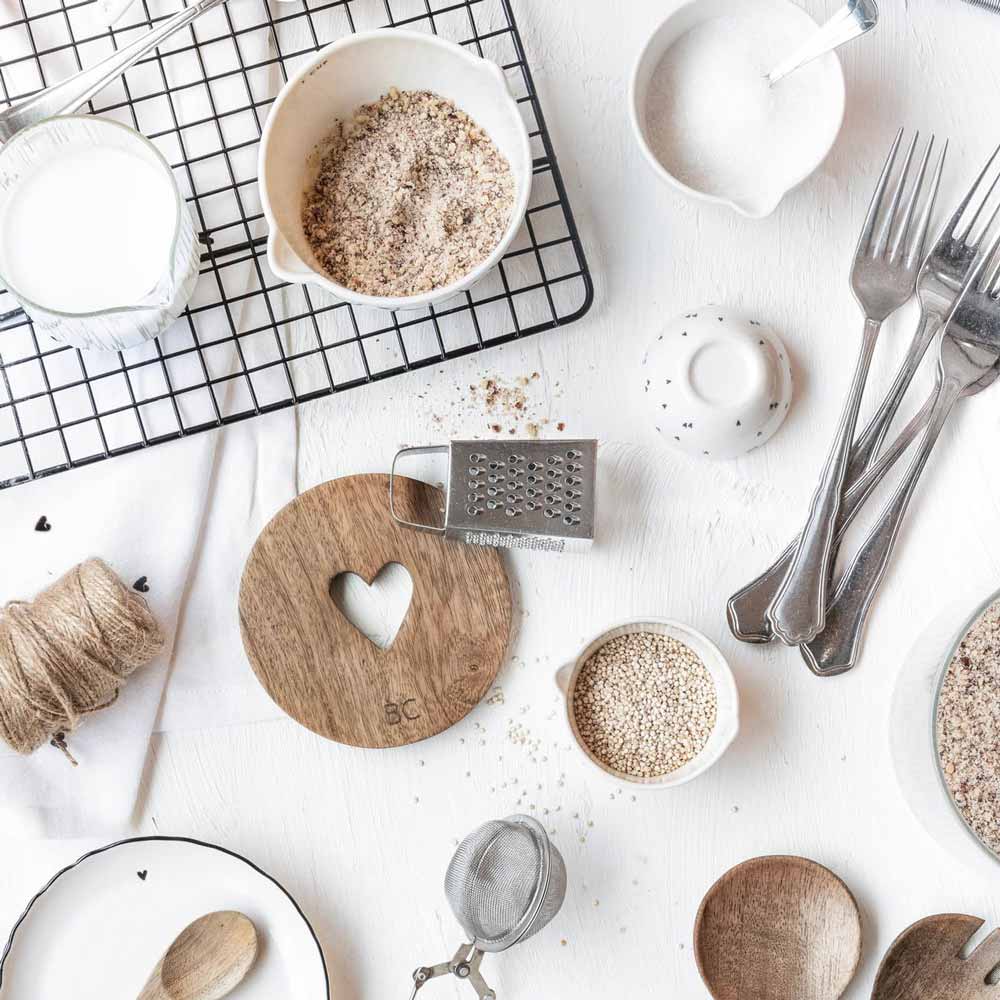 Auf einem Tisch sind eine kleine Reibe, Schüsseln mit Getreide, ein Glas Milch, verschiedene Utensilien, Schnur auf einem Kühlgitter und der kleine Holzuntersetzer „Holz Herz“ von Bastion Collections mit Herzausschnitt ausgestellt, alles auf einer weißen Oberfläche angeordnet.