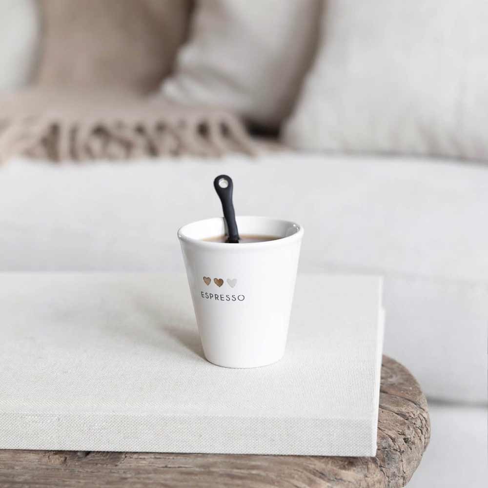 Eine wellenförmige Tasse von Bastion Collections – Löffel für Espresso, elegant mit der Aufschrift „ESPRESSO“ und drei Herzsymbolen beschriftet und mit einem schwarzen Löffel in der Hand, steht charmant auf einem weißen Buch auf einem Holztisch. Als Hintergrund dient ein beiges Sofa, das mit einer kuscheligen Decke geschmückt ist.