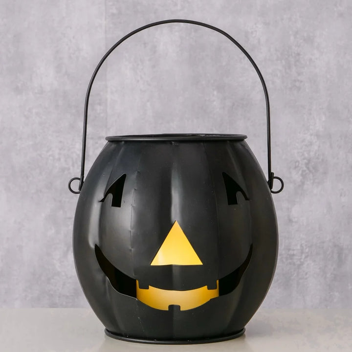 Das Boltze Halloween Windlicht Grimasi schwarzer Kürbis, eine schwarze Metalllaterne mit lächelndem Gesicht und Griff, leuchtet von innen, während es auf einer hellen Fläche vor einem grauen Hintergrund steht.