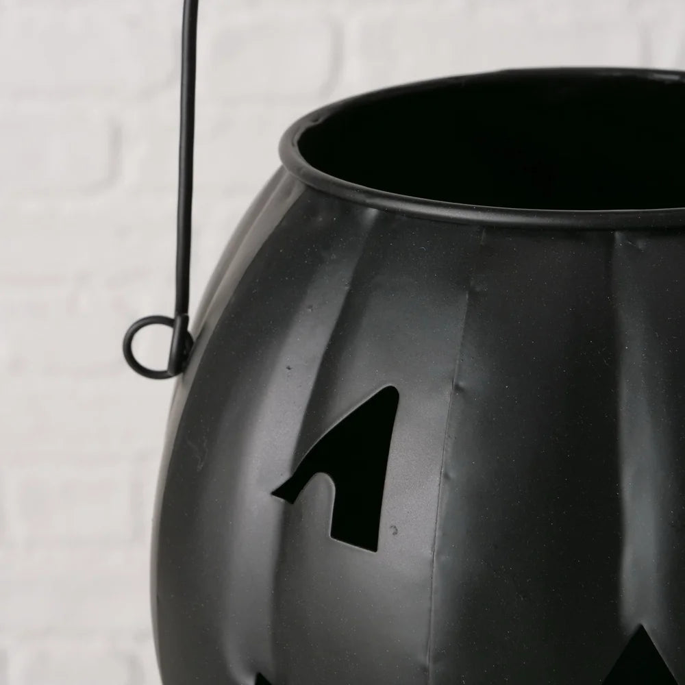 Nahaufnahme des Boltze Halloween Windlichts Grimasi schwarzer Kürbis, einer schwarzen Metalllaterne in Kürbisform mit ausgeschnittenem Gesicht und Griff, vor einem weißen Backsteinhintergrund.