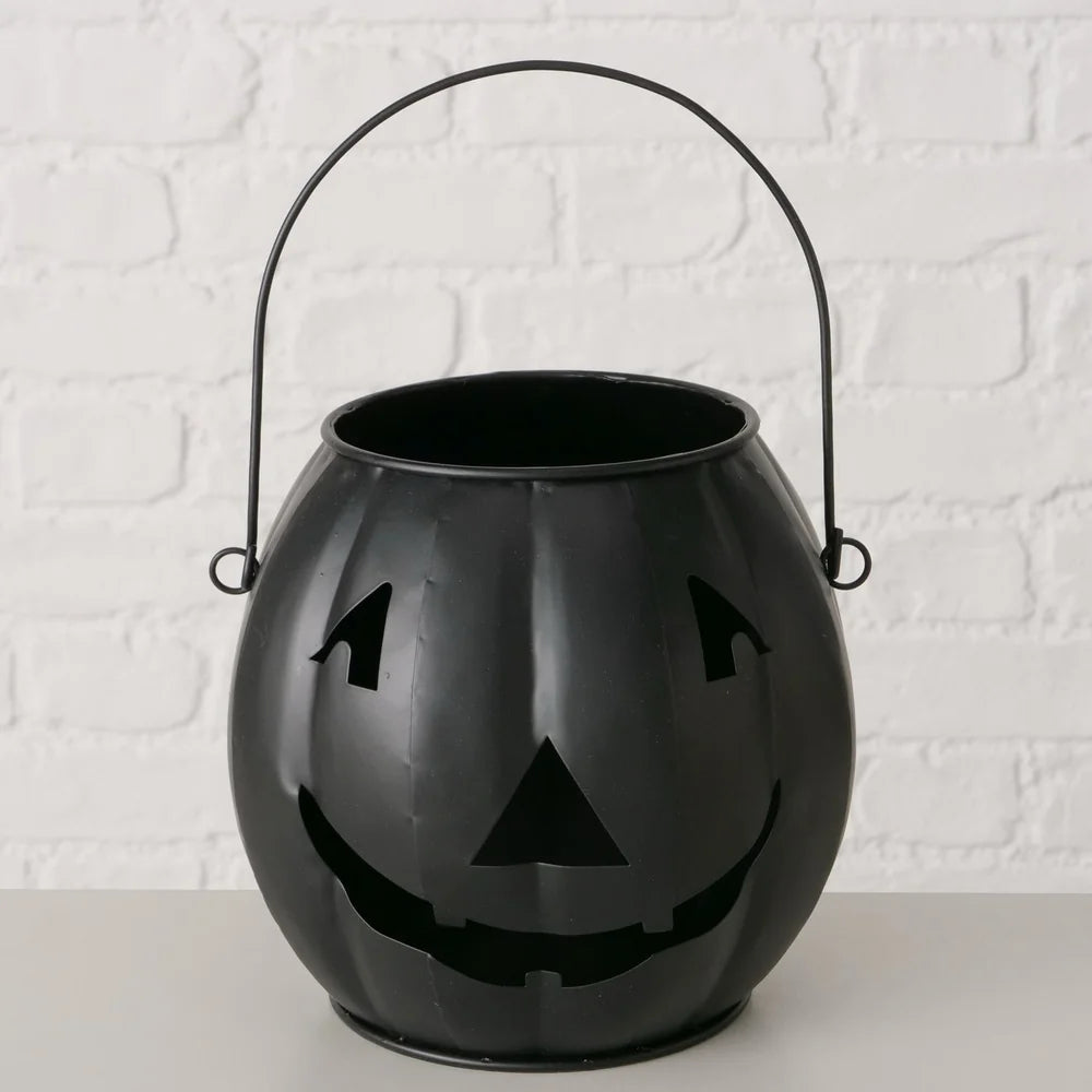 Das Boltze Halloween Windlicht Grimasi schwarzer Kürbis ist eine Kürbislaterne aus schwarzem Metall mit einem geschnitzten Kürbisgesicht und -griff, die auf einer hellen Fläche vor einer weißen Ziegelwand gezeigt wird.