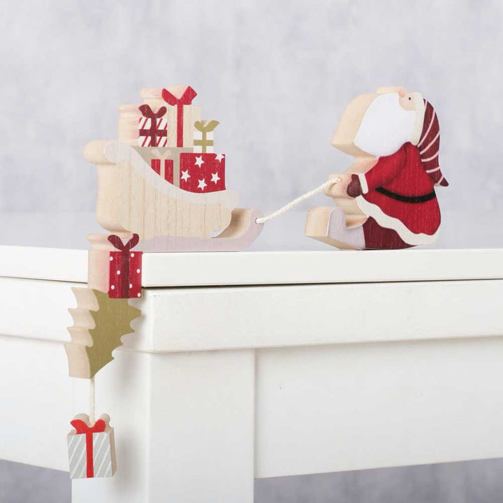 Boltzes Kantenhocker Weihnachtsmann mit Kutsche steht auf einem weißen Tisch und zeigt eine hölzerne Weihnachtsmannfigur, die einen Schlitten voller Geschenke zieht, über dessen Kante ein kleines Geschenk hängt.