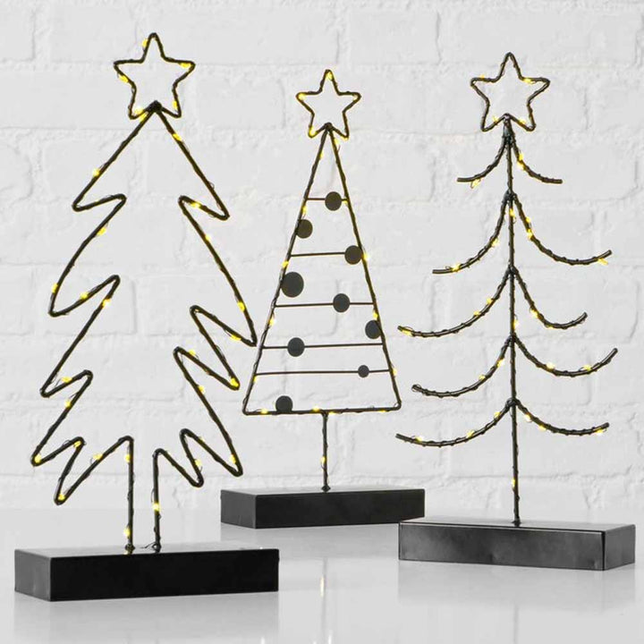 Drei metallene LED-Weihnachtsbaum-Silhouetten von Boltze, jeweils mit einem Stern versehen und mit gelben Lichterketten auf schwarzen Sockeln geschmückt, stehen vor einer weißen Backsteinwand.