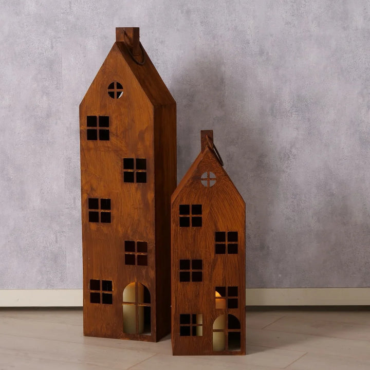 Das Boltze Laterne Alba Rost Deko Haus Mega 2er-Set zeigt zwei hölzerne Hauslaternen mit quadratischen Fenstern und bogenförmigen Türausschnitten, die auf einem gefliesten Boden vor einer strukturierten grauen Wand stehen.