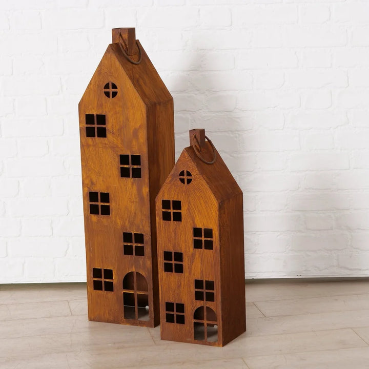 Das Boltze Laterne Alba Rost Deko Haus Mega 2er-Set besteht aus zwei hölzernen Hauslaternen mit ausgeschnittenen Fenstern und Türen, die auf einem hellen Boden vor einer weißen Backsteinwand stehen.