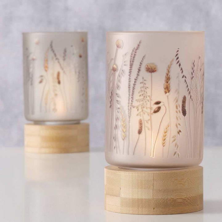 Zwei Kerzenhalter aus Milchglas „Boltze - Windlicht Jelena Floral auf Holzsockel“ mit aufwendigen botanischen Motiven und Holzsockeln, einer prominent im Vordergrund und der andere dezent im Hintergrund auf einer hellen Oberfläche.