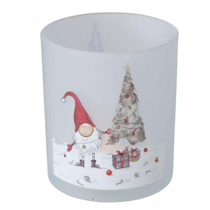 Ein Kerzenhalter aus Milchglas von Boltze aus der Windlicht Wichtel Bambi 2er-Set-Kollektion, der einen Gnom mit roter Mütze neben einem geschmückten Weihnachtsbaum und verpackten Geschenken vor einer schneebedeckten Kulisse zeigt.