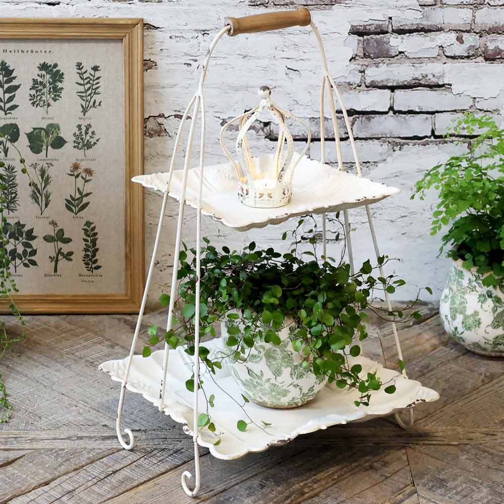 Zweistöckiger weißer Metallständer mit einer kleinen Krone als Ornament oben drauf. Auf der unteren Etage steht eine Topfpflanze mit grünem Blattwerk. Im Hintergrund sind ein gerahmter botanischer Druck und eine weitere Pflanze zu sehen. Chic Antique - Etagere mit Henkel