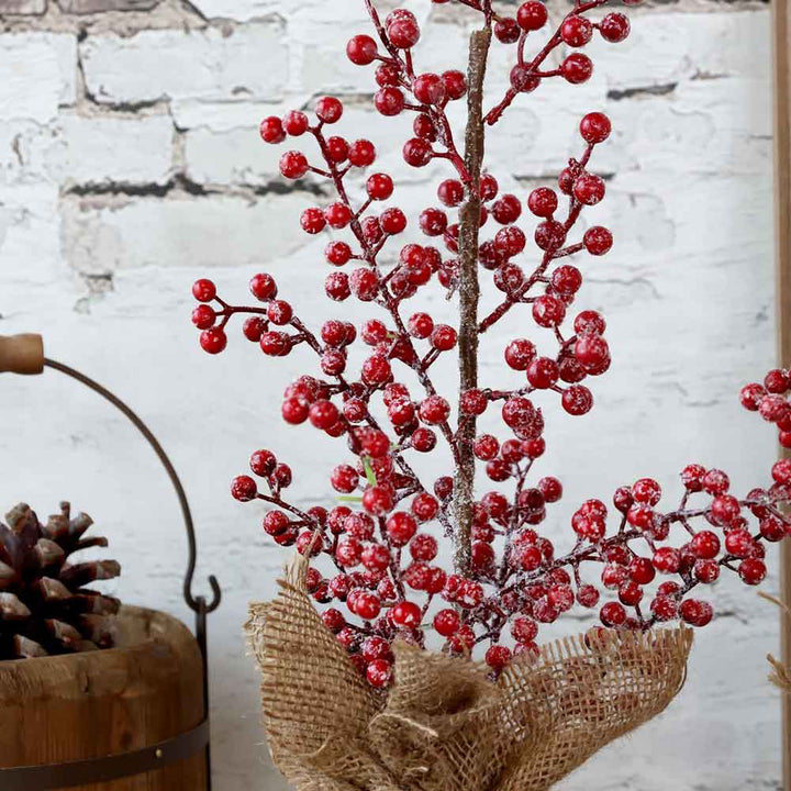 Ein Chic-Antique - Fleur Baum mit Beeren Höhe 40 cm wird elegant neben einem mit Tannenzapfen gefüllten Holzeimer vor einer weißen Backsteinwand präsentiert.